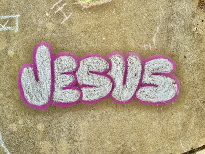 The image features the word 'JESUS' written in large, stylized graffiti letters. The letters are filled with a textured white or light gray color and are outlined with a bold pink or fuchsia color. The graffiti is on a concrete surface that has a weathered appearance and includes a few scattered and faintly chalked markings in various areas.