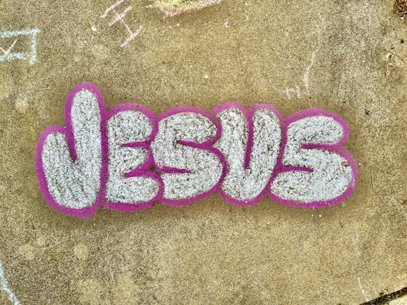 The image features the word 'JESUS' written in large, stylized graffiti letters. The letters are filled with a textured white or light gray color and are outlined with a bold pink or fuchsia color. The graffiti is on a concrete surface that has a weathered appearance and includes a few scattered and faintly chalked markings in various areas.