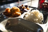 A metallic food tray divided into sections containing various foods. There is a portion of white rice in one section and a serving of fried food, possibly chicken or another type of breaded meat, in another. Another section contains a dish with dark-colored sauce. A person with chopsticks is visible in the background.