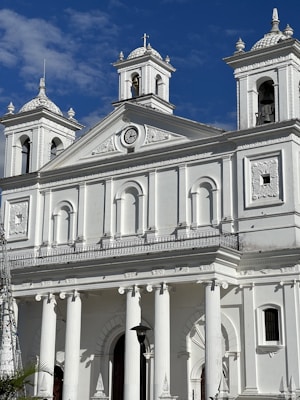 A grand, white neoclassical-style cathedral with tall columns and ornate architectural details. The facade features decorative sculptures and a prominent clock. Two bell towers rise on either side, each topped with a small dome.