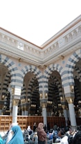 Several people are sitting and milling about in an architectural space featuring large, intricately designed arches with striped patterns. The arches are ornamented with gold and cream details. Shelving units line the back wall, and the people are dressed in various types of traditional clothing.