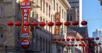A street scene in a bustling urban area with traditional red Chinese lanterns hanging across the street. Brick buildings with signs in both English and Chinese line the sides, including a prominent 'Eastern Bakery' sign. The architecture is a mix of classic and modern styles, with a tall modern building visible in the background.