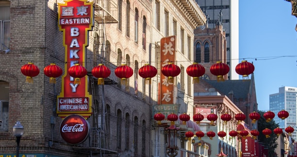 A street scene in a bustling urban area with traditional red Chinese lanterns hanging across the street. Brick buildings with signs in both English and Chinese line the sides, including a prominent 'Eastern Bakery' sign. The architecture is a mix of classic and modern styles, with a tall modern building visible in the background.