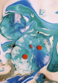 Abstract liquid art features swirling patterns of blue, green, and white with large and small spherical shapes and orange accents scattered throughout. The composition resembles a cosmic or underwater scene with fluid motion and dynamic contrasts.