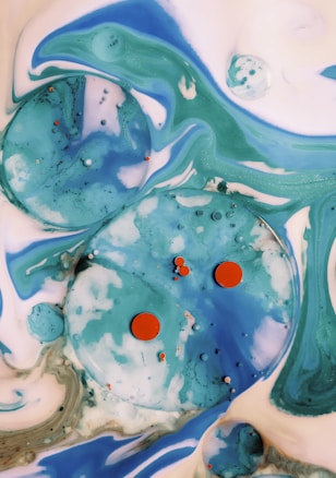 Abstract liquid art features swirling patterns of blue, green, and white with large and small spherical shapes and orange accents scattered throughout. The composition resembles a cosmic or underwater scene with fluid motion and dynamic contrasts.