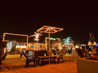 A nighttime beach scene featuring outdoor seating with various illuminated decorations and signs. Several palm trees and umbrellas are wrapped with lights, creating a festive atmosphere. Luminous signage for different places including 'GOA,' 'PARADISE,' and 'SEA TURTLE' can also be seen. There are chairs and tables placed in the sand, contributing to a casual and relaxed setting.