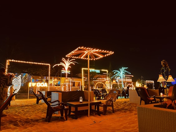 A nighttime beach scene featuring outdoor seating with various illuminated decorations and signs. Several palm trees and umbrellas are wrapped with lights, creating a festive atmosphere. Luminous signage for different places including 'GOA,' 'PARADISE,' and 'SEA TURTLE' can also be seen. There are chairs and tables placed in the sand, contributing to a casual and relaxed setting.