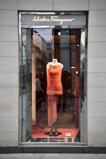 A storefront displays a vibrant orange dress on a mannequin. The dress features a fitted, sleeveless design and stands out against a neutral background. Reflective glass surfaces create subtle reflections of the street and surrounding architecture.