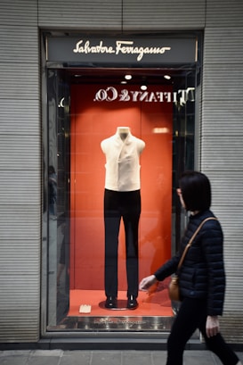 A store window display features a mannequin dressed in a white sleeveless top and black pants. The backdrop is a vibrant orange, and the brand name Salvatore Ferragamo appears above the display. A person walks past the store, wearing a black jacket and carrying a brown bag.