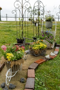 A charming garden display featuring decorative metal plant stands shaped like bicycles, each holding vibrant flowers in hues of yellow, pink, and purple. The garden is bordered by bricks and small decorative stone birds, with trellises supporting climbing plants. The setting is a lush green lawn under an overcast sky.