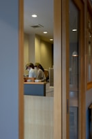 Two people wearing caps and uniforms appear to be working in a contemporary kitchen or cafe setting seen through a wooden-framed glass partition. The lighting is soft with ceiling lights providing a warm tone to the room.