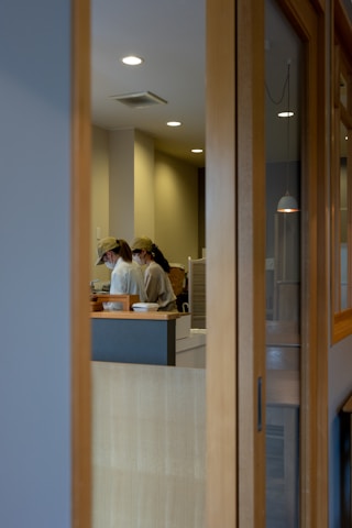 Two people wearing caps and uniforms appear to be working in a contemporary kitchen or cafe setting seen through a wooden-framed glass partition. The lighting is soft with ceiling lights providing a warm tone to the room.