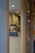 Two people wearing caps and uniforms appear to be working in a contemporary kitchen or cafe setting seen through a wooden-framed glass partition. The lighting is soft with ceiling lights providing a warm tone to the room.