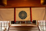 A traditional curtain with a blue chrysanthemum emblem hangs in front of a series of steps leading to an interior space. The setting suggests a cultural or historical significance, possibly in a temple or shrine. The curtain is bordered by red and is suspended under a wooden beam.