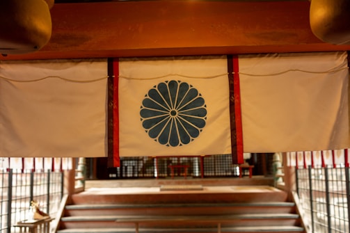 A traditional curtain with a blue chrysanthemum emblem hangs in front of a series of steps leading to an interior space. The setting suggests a cultural or historical significance, possibly in a temple or shrine. The curtain is bordered by red and is suspended under a wooden beam.
