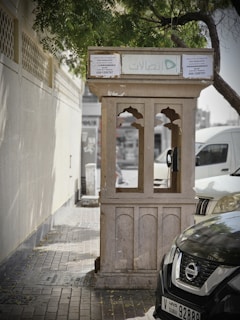 A weathered public telephone booth stands on a paved sidewalk next to a wall and parked cars. The booth, with arched windows and a flat roof, is partly shaded by tree branches overhead. Text in Arabic and English is visible on the signage, and a green logo is prominently displayed. The street scene suggests a quiet, urban area.