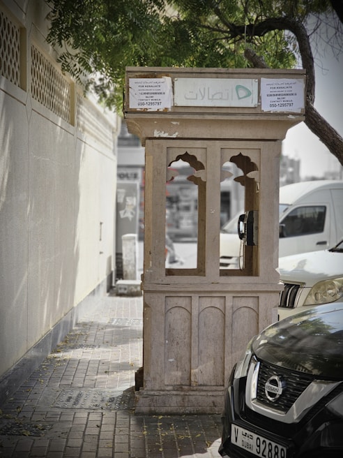 A weathered public telephone booth stands on a paved sidewalk next to a wall and parked cars. The booth, with arched windows and a flat roof, is partly shaded by tree branches overhead. Text in Arabic and English is visible on the signage, and a green logo is prominently displayed. The street scene suggests a quiet, urban area.