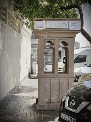 A weathered public telephone booth stands on a paved sidewalk next to a wall and parked cars. The booth, with arched windows and a flat roof, is partly shaded by tree branches overhead. Text in Arabic and English is visible on the signage, and a green logo is prominently displayed. The street scene suggests a quiet, urban area.