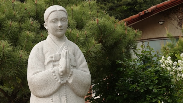 A stone statue with hands clasped in prayer stands amidst lush greenery, set against a backdrop of a tiled-roof building and pine trees.