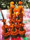 Several traditional floral arrangements are composed of vibrant orange marigold flowers. The flowers are intricately placed around conical green leaves. Incense sticks are inserted vertically into the arrangements. These items rest on a colorful fabric with various patterns and shades of red.