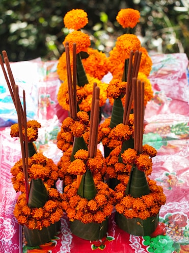 Several traditional floral arrangements are composed of vibrant orange marigold flowers. The flowers are intricately placed around conical green leaves. Incense sticks are inserted vertically into the arrangements. These items rest on a colorful fabric with various patterns and shades of red.