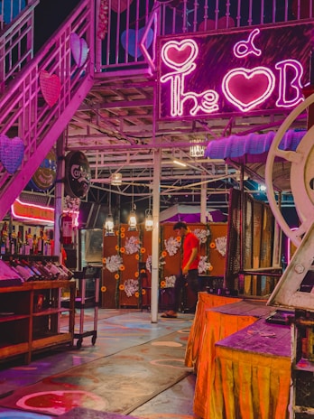 A vibrant indoor bar environment with neon pink lighting. The scene includes a staircase decorated with heart shapes, a neon sign, and a bar area stocked with various bottles. Hanging lights illuminate the space featuring a colorful floor with circular patterns. A person in a red shirt is walking by a floral-decorated wall.