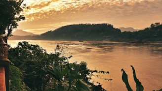 A tranquil riverside scene in Thailand at sunset, with soft golden light reflecting on calm water and lush greenery.