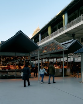 A spacious outdoor market with people walking and shopping. The market is covered with modern canopy structures, and various stalls are visible displaying an array of goods. The architectural design features on the upper level with decorative railings and drapes.