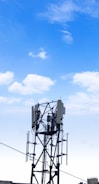 A cellular tower stands against a clear blue sky with scattered clouds. The structure features antennas and cables, characteristic of telecommunications equipment. Its metallic framework rises vertically with several extensions and components.