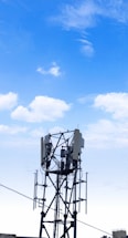 A cellular tower stands against a clear blue sky with scattered clouds. The structure features antennas and cables, characteristic of telecommunications equipment. Its metallic framework rises vertically with several extensions and components.