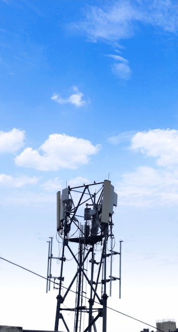 A cellular tower stands against a clear blue sky with scattered clouds. The structure features antennas and cables, characteristic of telecommunications equipment. Its metallic framework rises vertically with several extensions and components.