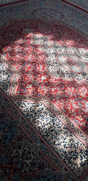 Intricate Persian carpet featuring a rich pattern of floral and geometric designs. The carpet predominantly showcases red and blue hues with detailed ornamentation in white and gold tones. Natural sunlight casts soft shadows across the surface, enhancing the texture and colors.