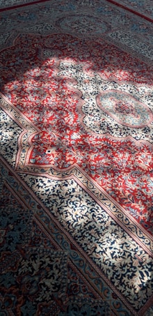 Intricate Persian carpet featuring a rich pattern of floral and geometric designs. The carpet predominantly showcases red and blue hues with detailed ornamentation in white and gold tones. Natural sunlight casts soft shadows across the surface, enhancing the texture and colors.