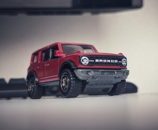A miniature red toy truck with prominent details is positioned on a flat surface. The model features bold lettering on the front grille, large tires, and a rugged design. The background is slightly blurred, focusing attention on the toy vehicle.