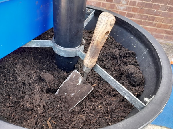 A trowel with a wooden handle is placed in a container filled with rich, dark soil. The container is made of metal and has a central black plastic support column. Behind the container, there is a brick wall.