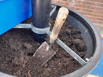 A trowel with a wooden handle is placed in a container filled with rich, dark soil. The container is made of metal and has a central black plastic support column. Behind the container, there is a brick wall.