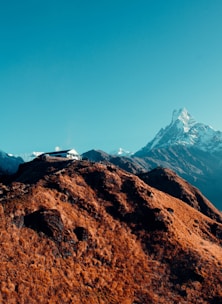 A solitary house is perched on a rugged, brown hillside with a sharp, snow-covered mountain peak towering in the background. The sky is clear and blue, contrasting with the earthy tones of the hill and the icy whites of the mountain.