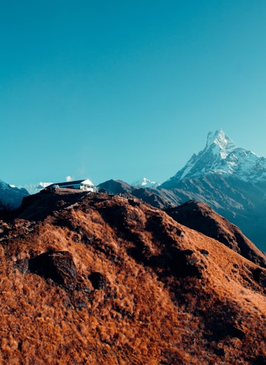 A solitary house is perched on a rugged, brown hillside with a sharp, snow-covered mountain peak towering in the background. The sky is clear and blue, contrasting with the earthy tones of the hill and the icy whites of the mountain.