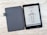 An e-reader with a black cover is open on a textured light-colored surface. The screen displays information in a foreign language with an illustration of a boat under the title 'KON-TIKI-3.' A series of specifications and icons is listed below.