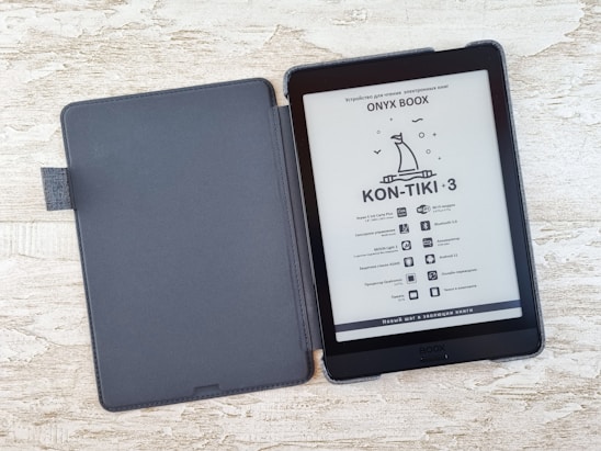 An e-reader with a black cover is open on a textured light-colored surface. The screen displays information in a foreign language with an illustration of a boat under the title 'KON-TIKI-3.' A series of specifications and icons is listed below.