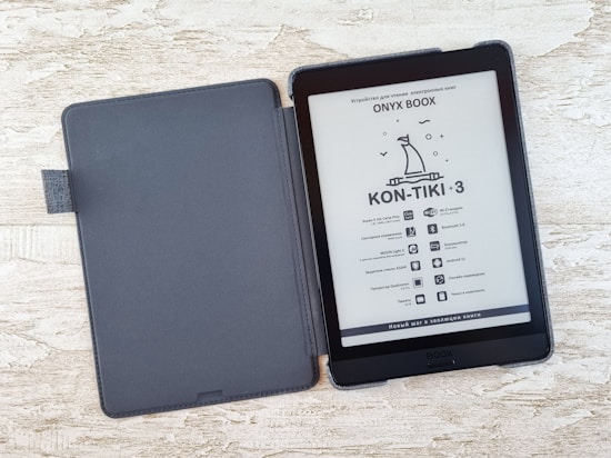 An e-reader with a black cover is open on a textured light-colored surface. The screen displays information in a foreign language with an illustration of a boat under the title 'KON-TIKI-3.' A series of specifications and icons is listed below.