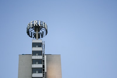 A tall building with a prominent sign displaying the word 'BERLIN' in large letters at the top. The sign is mounted on a circular framework above the structure. The building has a modern design with a series of windows aligned vertically and an external ladder on one side. The sky in the background is clear and blue.