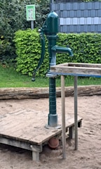 A green manual water pump is mounted on a wooden platform surrounded by sand. There is a lush hedge in the background and a sign with instructions or warnings. The water pump has a metal lever and spout.