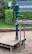 A green manual water pump is mounted on a wooden platform surrounded by sand. There is a lush hedge in the background and a sign with instructions or warnings. The water pump has a metal lever and spout.