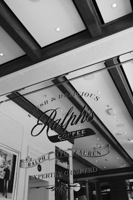A stylish black and white image featuring the interior of a coffee shop. The ceiling has wooden beams and recessed lighting, adding an elegant touch. A transparent glass displays the logo 'Ralph's Coffee Ralph Lauren' accompanied by phrases like 'Fresh & Delicious' and 'Expertly Crafted.'