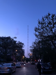 A street scene at dusk with a tall radio tower in the center, surrounded by trees and buildings. Streetlights are on, illuminating parked cars along the sides of the road, and a person is seen riding a motorcycle. The sky is a deep blue, adding to the serene atmosphere.