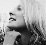 A black and white profile portrait of a person gazing upwards thoughtfully, with one hand resting under their chin. Their hair is light-colored and slightly tousled. In the background, blurry trees add depth to the composition.