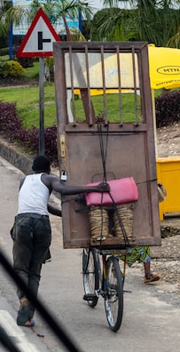 A person is pushing a bicycle loaded with a large wooden door, a pink container, and a bundle of straw. There is a road sign in the background, and a bright yellow umbrella partly covers another individual.
