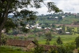 A rural landscape with scattered houses and abundant greenery. The scene is framed by trees in the foreground, offering a view of the lush countryside. Rolling hills and small dwellings composed of earthen roofs create a picturesque village setting under a cloudy sky.