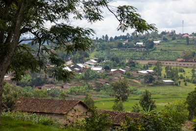 A rural landscape with scattered houses and abundant greenery. The scene is framed by trees in the foreground, offering a view of the lush countryside. Rolling hills and small dwellings composed of earthen roofs create a picturesque village setting under a cloudy sky.
